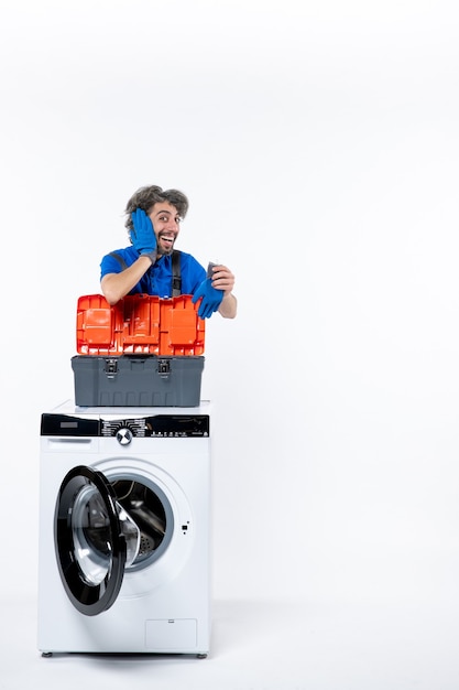 Front view elated repairman putting hand to his ear behind washing machine on white space