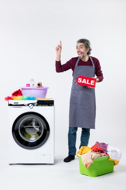 Front view elated young guy in apron holding up sale sign standing near washer on white background