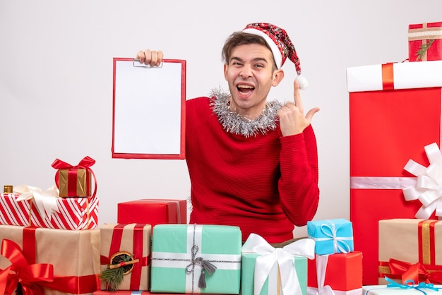 Front view elated young man holding clipboard sitting around xmas gifts