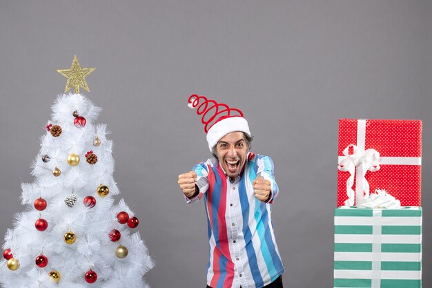 Вид спереди приподнятого молодого человека в новогодней шапке со спиральной пружиной, показывающей свое счастье возле белой рождественской елки