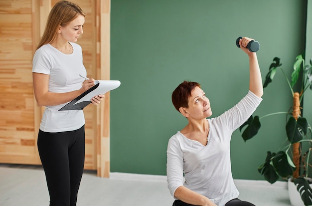 Front view of elder woman in covid recovery doing physical exercises with dumbbell while nurse checks