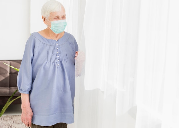 Free photo front view of elder woman looking through the window while wearing medical mask