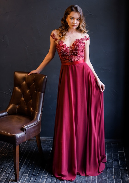 Front view of elegant girl in a red dress leans on a chair and looks at the camera