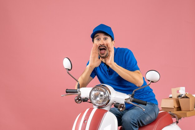 Front view of emotional courier guy wearing hat sitting on scooter and calling someone on pastel peach background