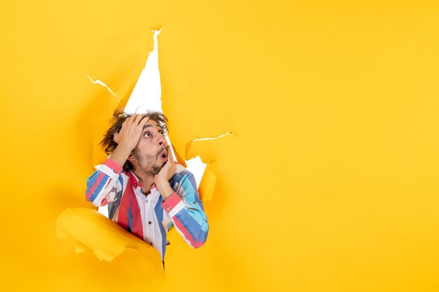 Front view of an emotional and crazy exhausted young guy looking up through a torn hole in yellow paper