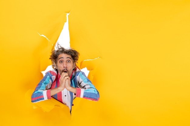 Free photo front view of an emotional and exhausted young man through a torn hole in yellow paper