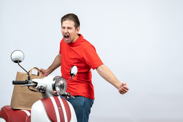 Front view of emotional nervous delivery guy in red uniform standing near scooter on white background