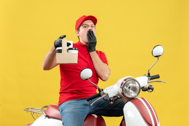 Front view of emotional shocked courier man wearing red blouse and hat gloves in medical mask delivering order sitting on scooter holding orders