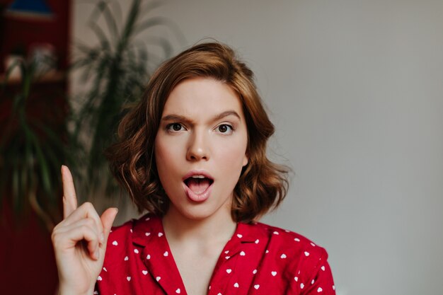 Front view of emotional woman in sleepwear showing idea sign. Indoor shot of curly woman in red pajama gesturing at home.