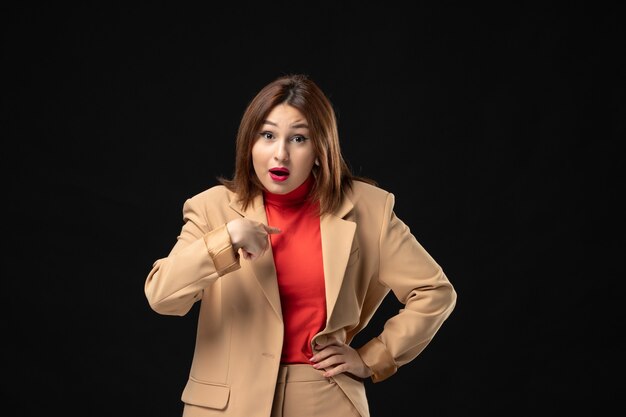 Front view of emotional young lady in a light brown suit pointing herself on dark