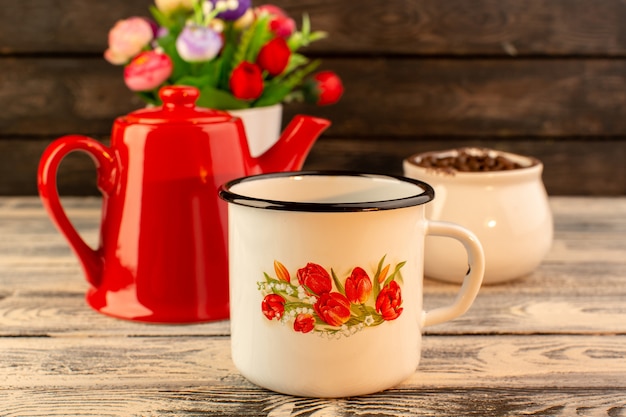 Front view of empty cup with red kettle brown coffee seeds and flowers on the wooden desk