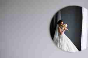 Free photo front view of empty grey wall and circle mirror on which is reflected attractive bride in wedding gown which raising up bouquet