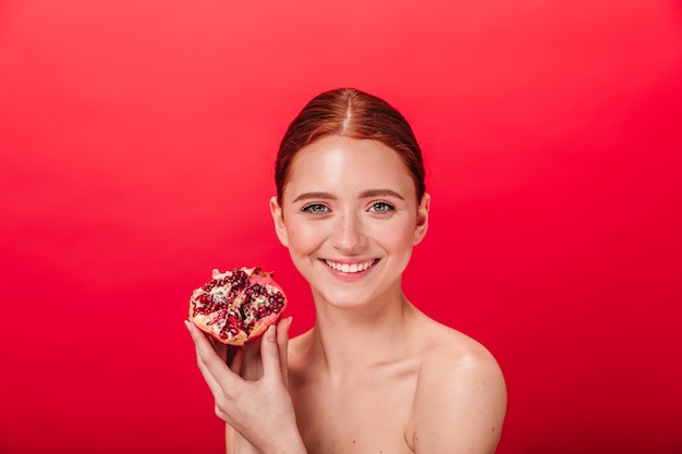 Vista frontale della donna incantevole che sorride e che tiene granato succoso. studio shot di spensierata ragazza allo zenzero con melograno.