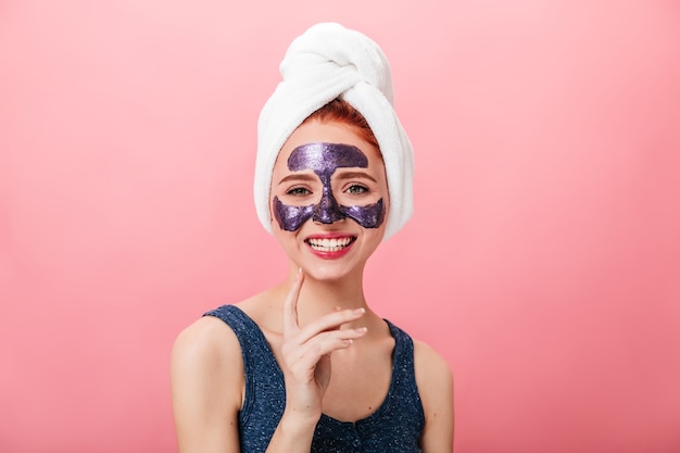 ピンクの背景で笑っているフェイスマスクを持つ魅惑的な女性の正面図。スパトリートメントをしている頭にタオルで至福の少女のスタジオショット。