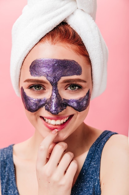 Front view of excited girl having fun during spa treatment. Studio shot of happy european woman with face mask smiling on pink background.