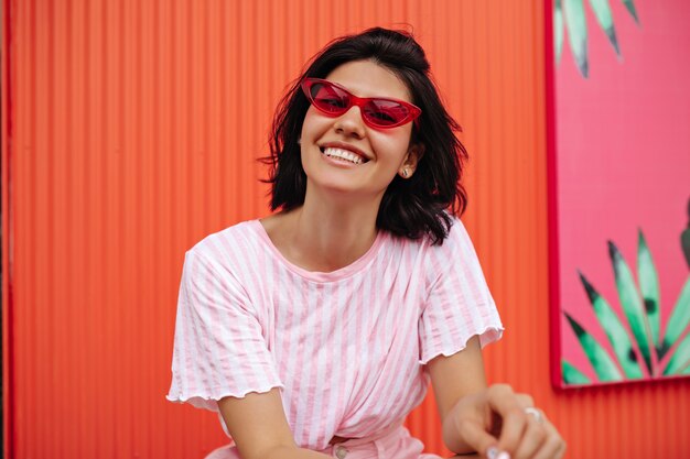 Front view of excited woman in striped t-shirt. Outdoor shot of laughing tanned woman in pink sunglasses.