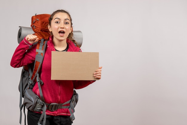 Front view of excited young traveller with big backpack holding cardboard on grey wall