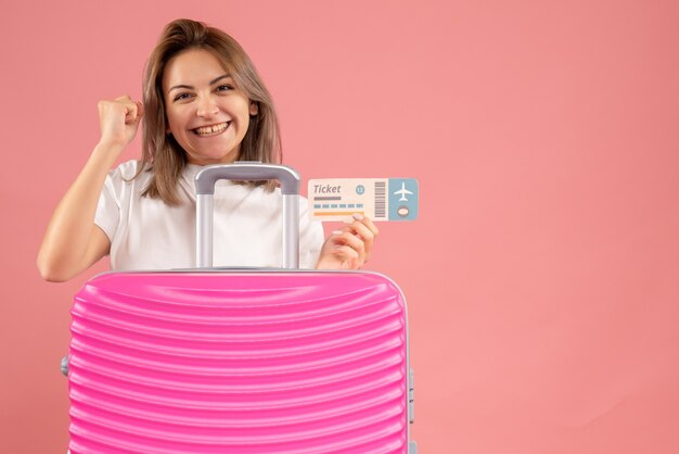 Front view of excited young woman holding ticket behind pink suitcase