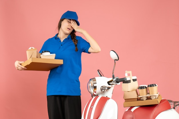 Front view of exhausted courier girl standing next to motorcycle holding coffee and small cakes on pastel peach color background