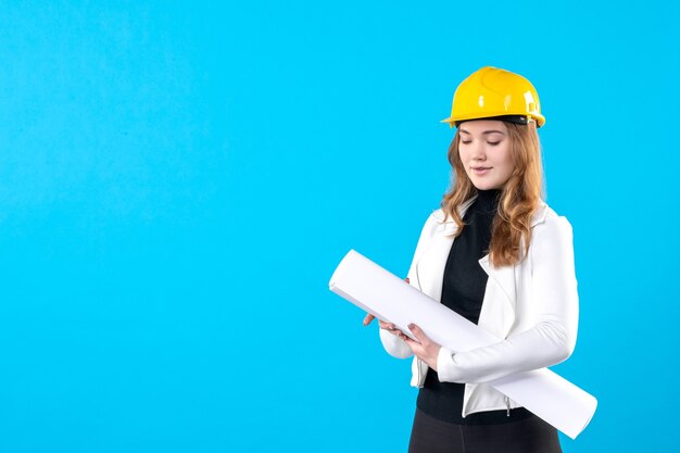 Front view female architect in yellow helmet holding plan on blue