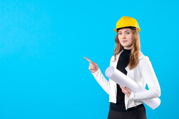 Front view female architect in yellow helmet holding plan on blue