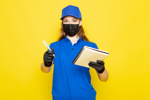 Free photo a front view female attractive courier in blue polo shirt blue cap and jeans in black gloves black protective mask holding pen and notebook on the yellow background food service job