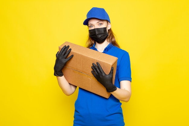 A front view female attractive courier in blue polo shirt blue cap and jeans holding package in black gloves black protective mask posing on the yellow background food service job