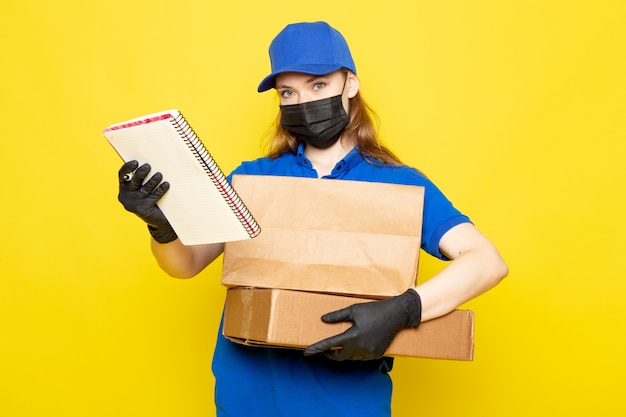 A front view female attractive courier in blue polo shirt blue cap and jeans holding package in black gloves black protective mask posing on the yellow background food service job