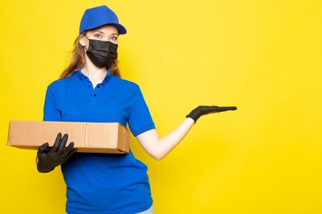 青いポロシャツブルーキャップとジーンズを保持している正面の女性の魅力的な宅配便黒い手袋黒い防護マスクでパッケージを保持している黄色の背景のフードサービスの仕事でポーズ