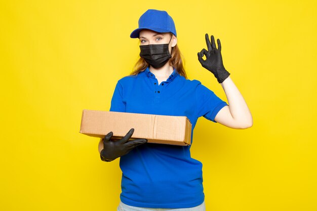 青いポロシャツブルーキャップとジーンズを保持している正面の女性の魅力的な宅配便は黄色の背景のフードサービスの仕事で黒い手袋黒い防護マスクでパッケージを保持しています。