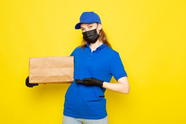 A front view female attractive courier in blue polo shirt blue cap and jeans holding package in black gloves black protective mask on the yellow background food service job