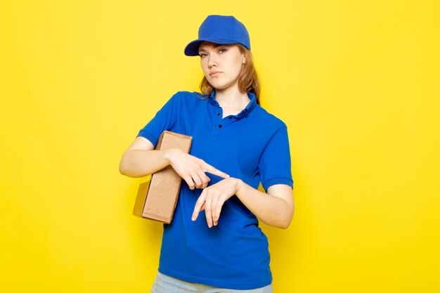 A front view female attractive courier in blue polo shirt blue cap and jeans holding package touching her wrist on the yellow background food service job