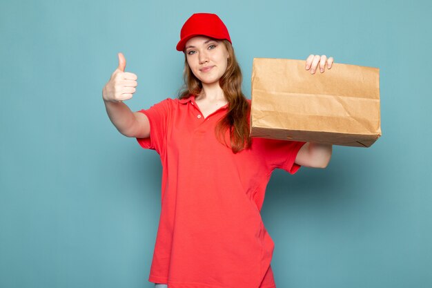 A front view female attractive courier in red polo shirt red cap holding brown package smiling on the blue background food service job