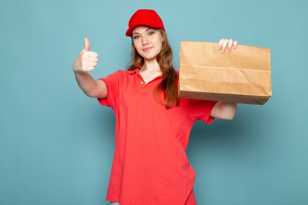 青い背景のフードサービスの仕事に笑みを浮かべて茶色のパッケージを保持している赤いポロシャツの赤い帽子の正面の女性の魅力的な宅配便