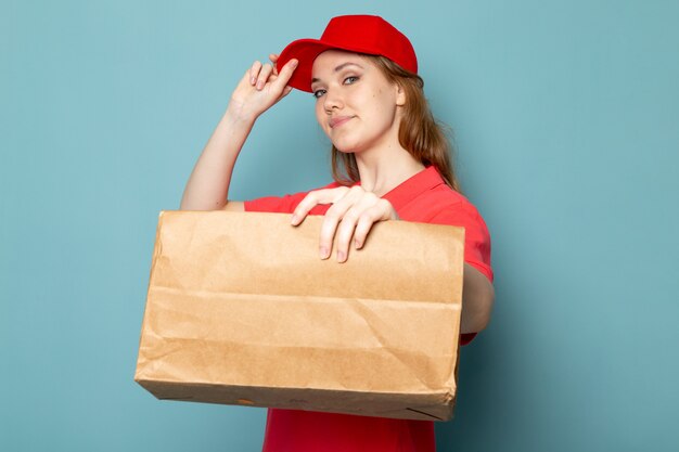 파란색 배경 음식 서비스 작업에 포즈 웃고 갈색 패키지를 들고 빨간 폴로 셔츠 빨간 모자에 전면보기 여성 매력적인 택배