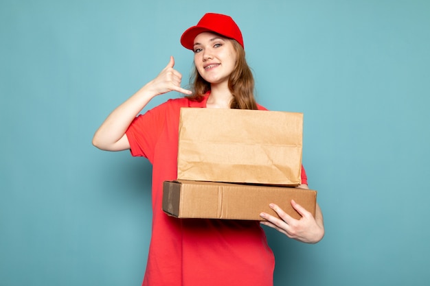 A front view female attractive courier in red polo shirt red cap holding brown package using her imaginary phone smiling on the blue background food service job