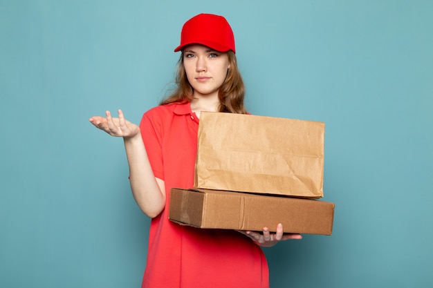 青い背景のフードサービスの仕事に茶色のパッケージを保持している赤いポロシャツの赤い帽子の正面の女性の魅力的な宅配便