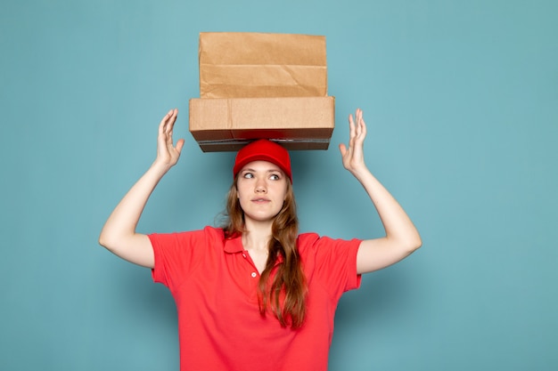 A front view female attractive courier in red polo shirt red cap holding brown packages above her head posing on the blue background food service job