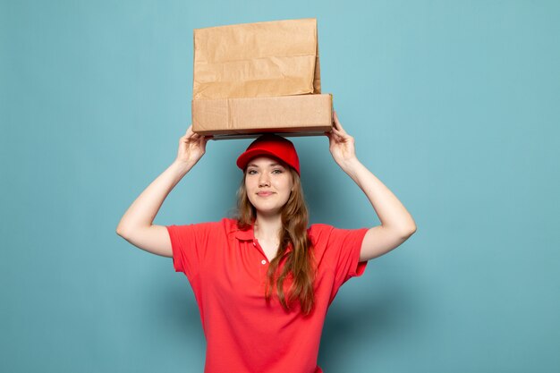 A front view female attractive courier in red polo shirt red cap holding brown packages smiling posing above her head on the blue background food service job