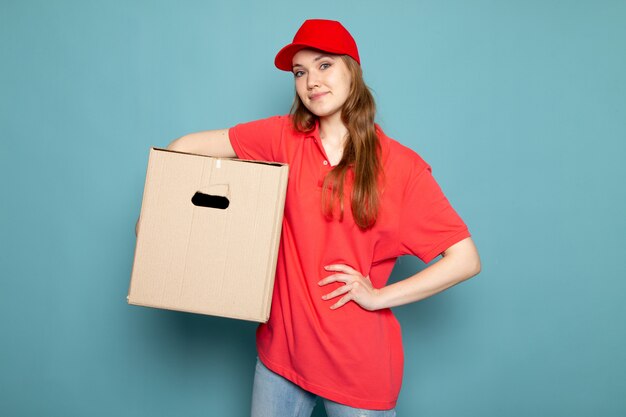 A front view female attractive courier in red polo shirt red cap and jeans holding box posing on the blue background food service job