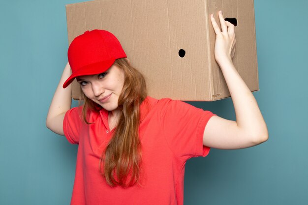 A front view female attractive courier in red polo shirt red cap and jeans holding package box posing on the blue background food service job