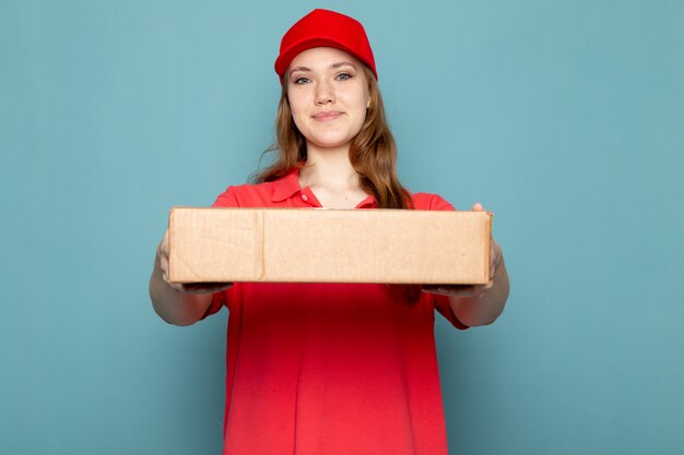 Вид спереди женский привлекательный курьер в красной рубашке поло, красной кепке и джинсах держит пакет позирует на синем фоне работы общественного питания