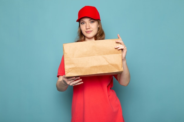 Un corriere attraente femminile di vista frontale in spiritello malevolo rosso della camicia di polo e jeans che tengono pacchetto che posa sorridere sul lavoro blu di servizio ristoro del fondo