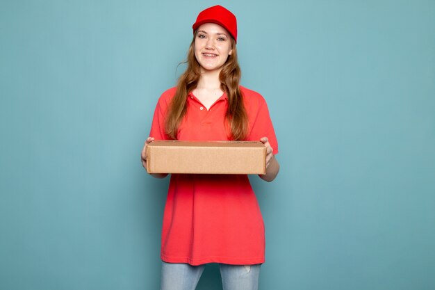 A front view female attractive courier in red polo shirt red cap and jeans holding package posing smiling on the blue background food service job