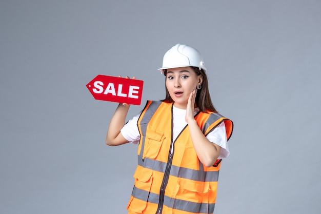 Front view of female builder holding red sale board on gray wall