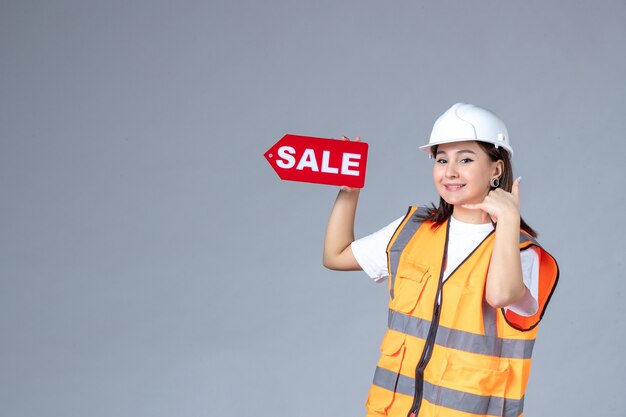 Вид спереди женщины-строителя, держащей красную доску продажи на серой стене