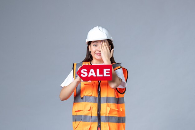 Front view of female builder holding red sale board on gray wall