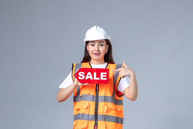 Front view of female builder holding red sale board on gray wall