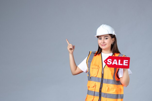 Front view of female builder holding red sale board on gray wall