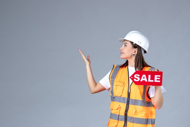 Front view of female builder holding red sale board on gray wall